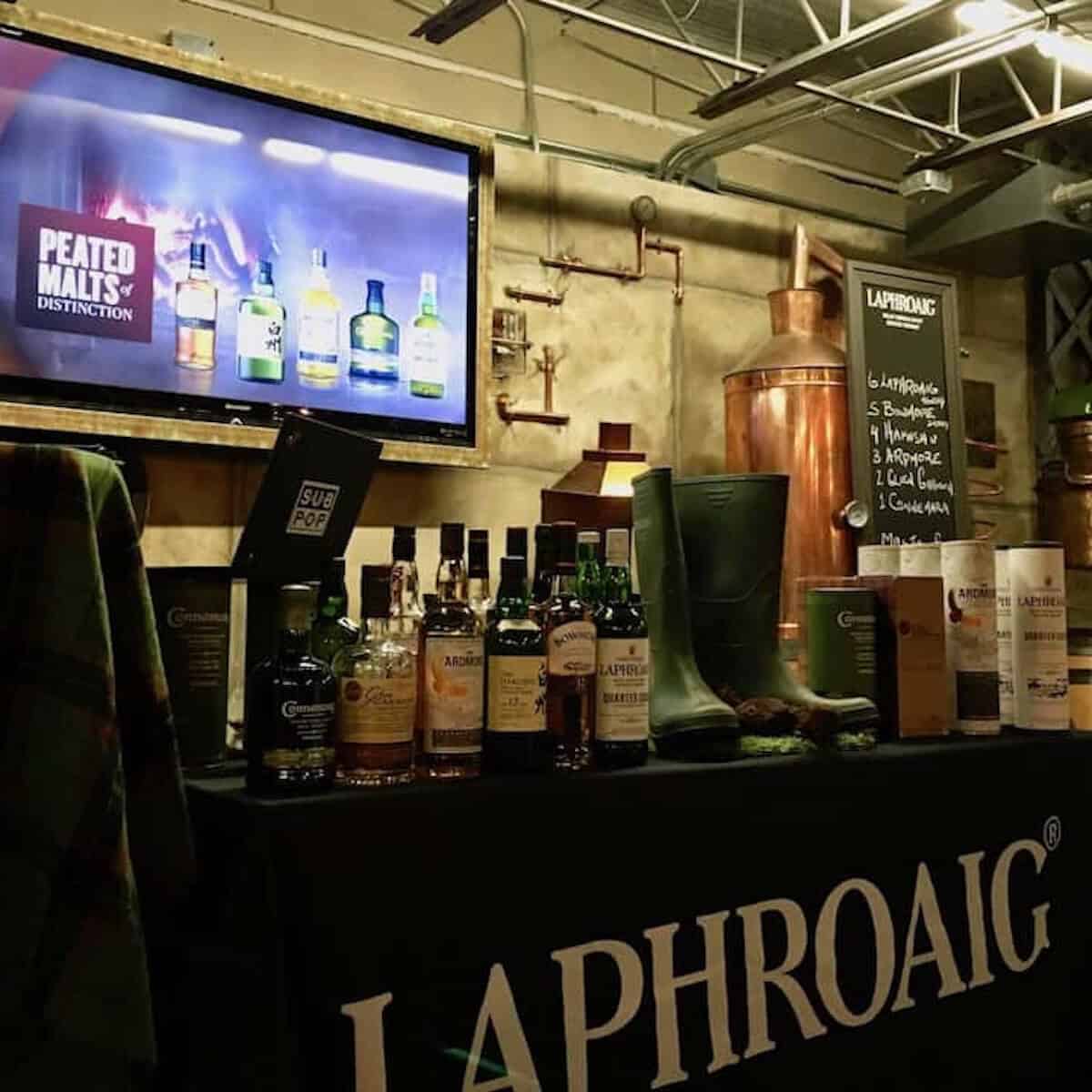 Table with whisky bottles, boots and a tartan with a tv display in background.