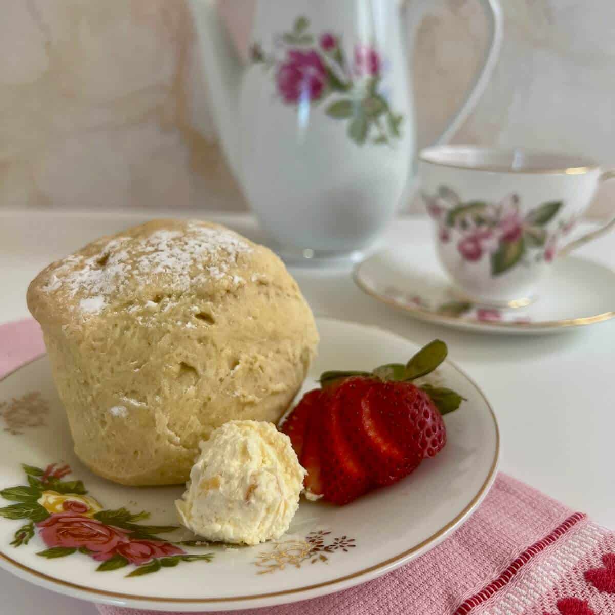 Clotted cream alongside a scone & cut strawberry on a flowered plate with teapot & teacup behind.