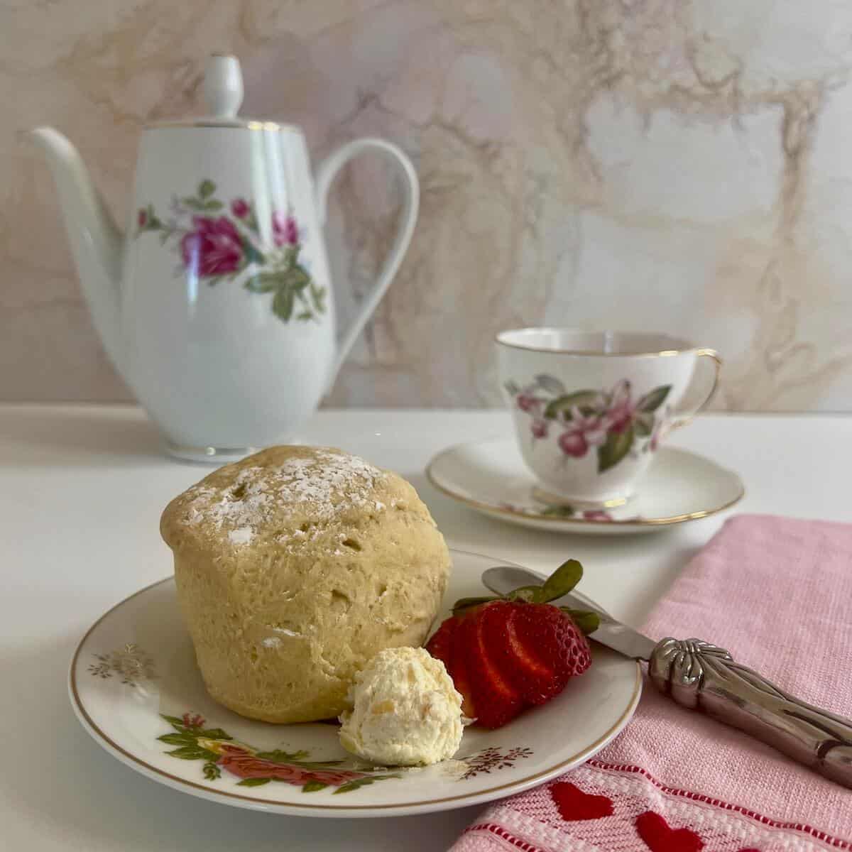 Clotted cream alongside a scone & cut strawberry on a flowered plate with teapot & teacup behind.