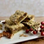 Pecan bars stacked on a white plate surrounded by pecans, brown sugar, and red berries.