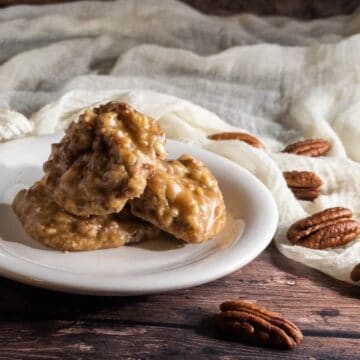 Pecan pralines stacked on a white plate with pecans & ivory scarf behind.