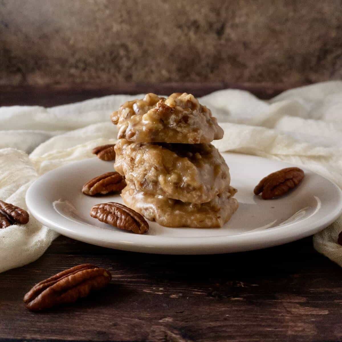 Pecan pralines stacked on a white plate with pecans & ivory scarf behind.