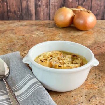 French Onion Soup in a bowl next to a spoon on a grey napkin with onions behind.