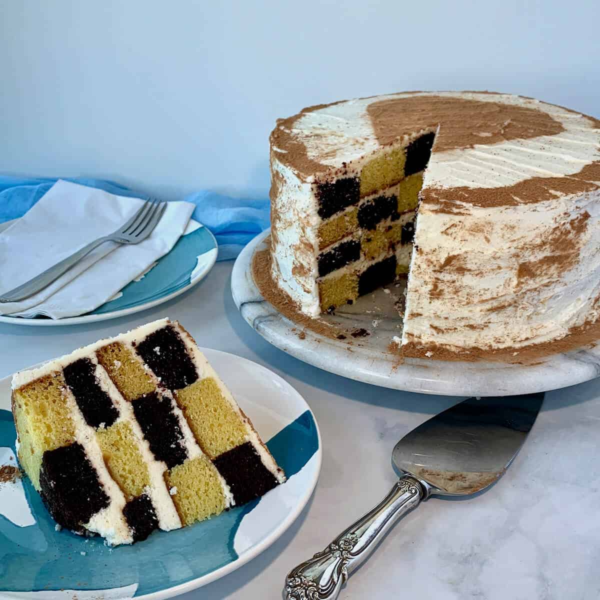 Checkerboard cake slice on a blue & white plate next to a cake serve with cake on plate and fork in background.