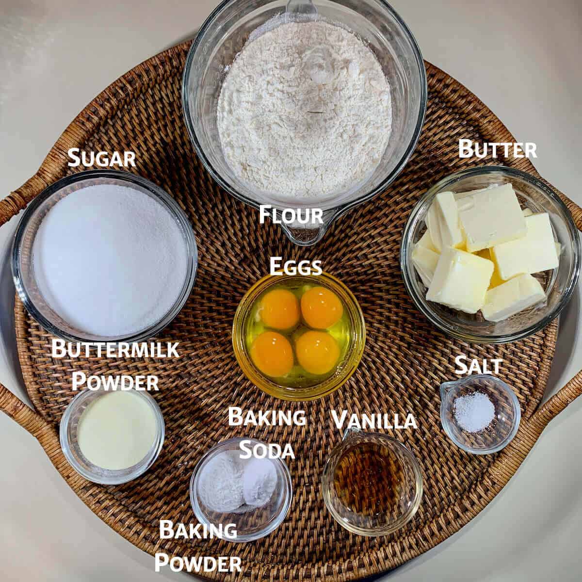 Vanilla cake ingredients portioned into glass bowls on a wooden tray from overhead.