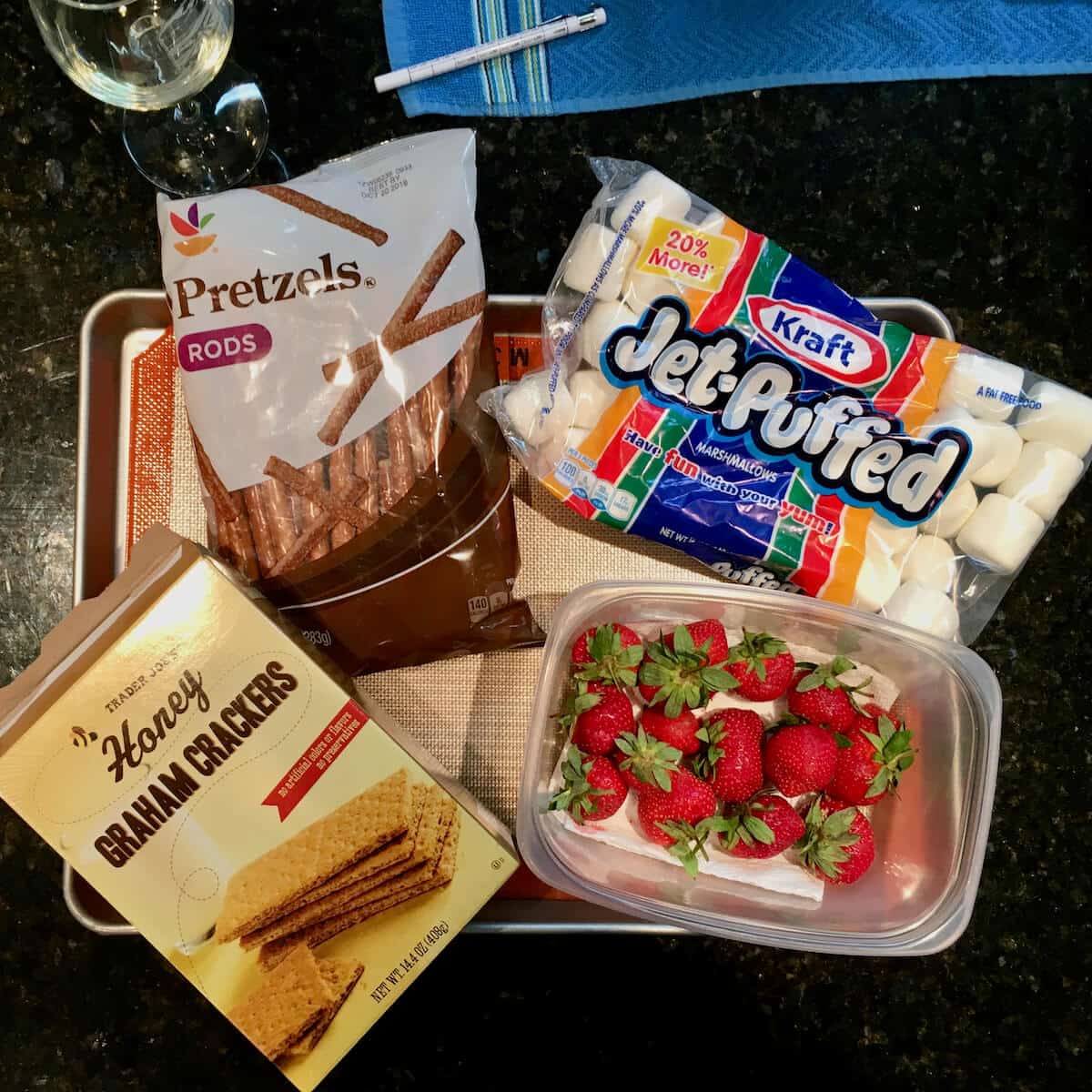 Pretzels, marshmallows, graham crackers and strawberries on a baking pan set on a dark marble counter.