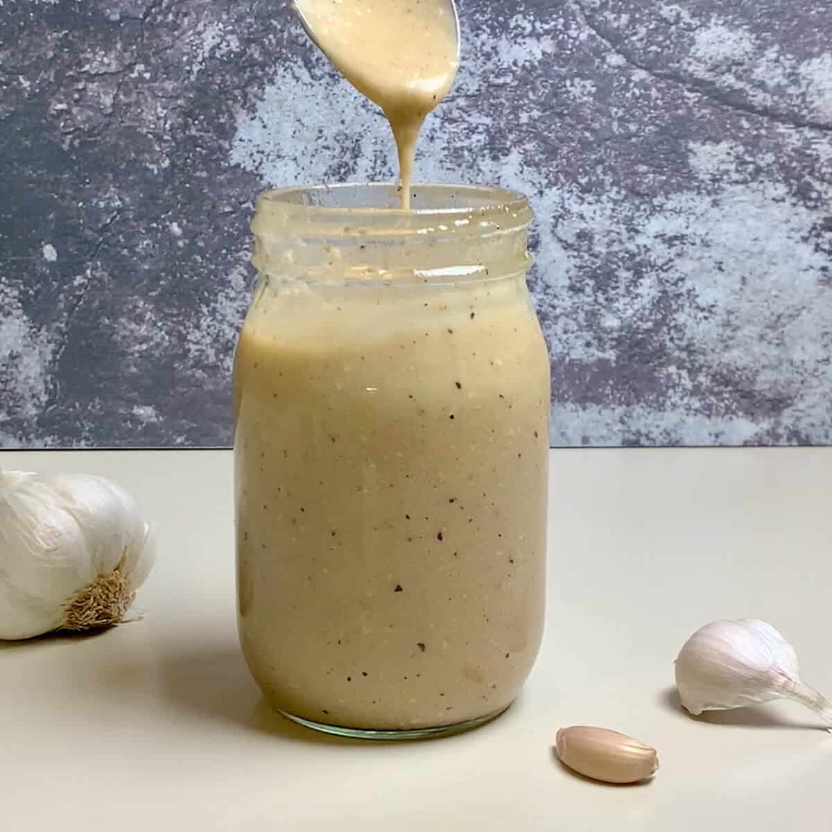 Spoon dripping Caesar dressing into a glass jar with garlic on a counter.