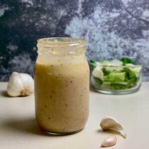 Jar of Caesar salad dressing on a counter with garlic & green salad.