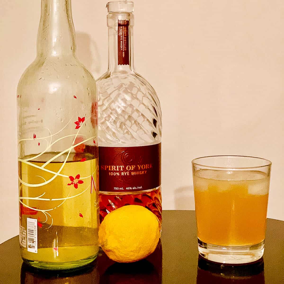 Limoncello Rye Sour in a glass with a lemon and bottles of Limoncello & Rye Whiskey on a black table.