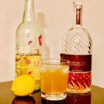 Limoncello Rye Sour in a glass with a lemon and bottles of Limoncello & Rye Whiskey.