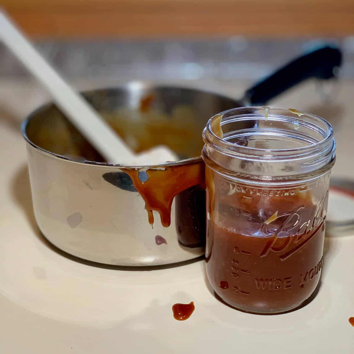 Caramel sauce in jar in front of a saucepan on a counter.