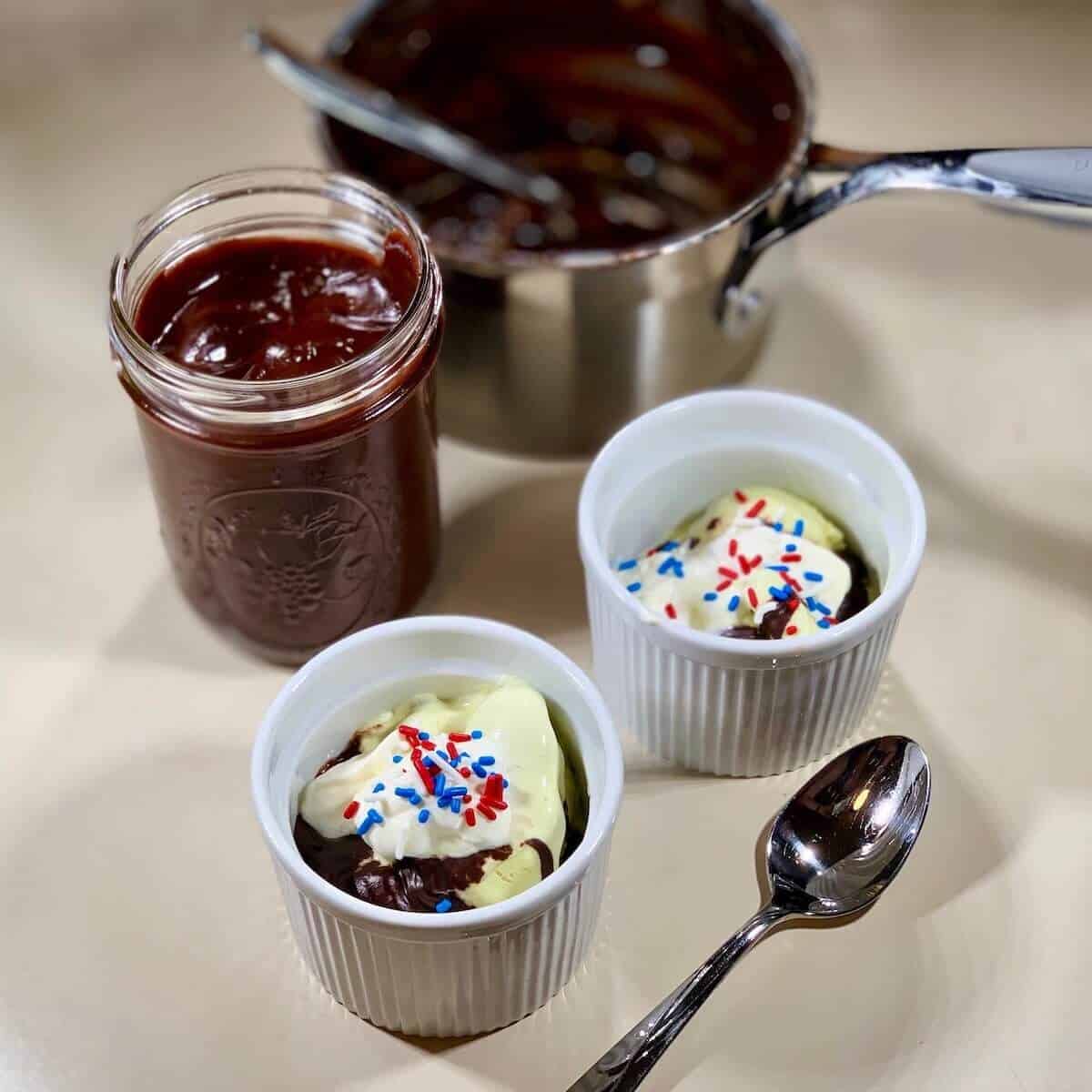 2 hot fudge sundaes with a spoon, jar of sauce, and saucepan.