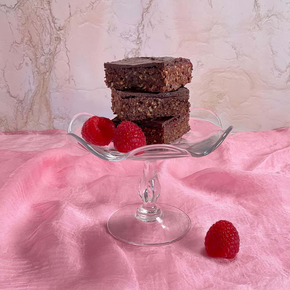 Three Chocolate Raspberry Brownies & fresh raspberries stacked on a glass stand on a pink scarf.