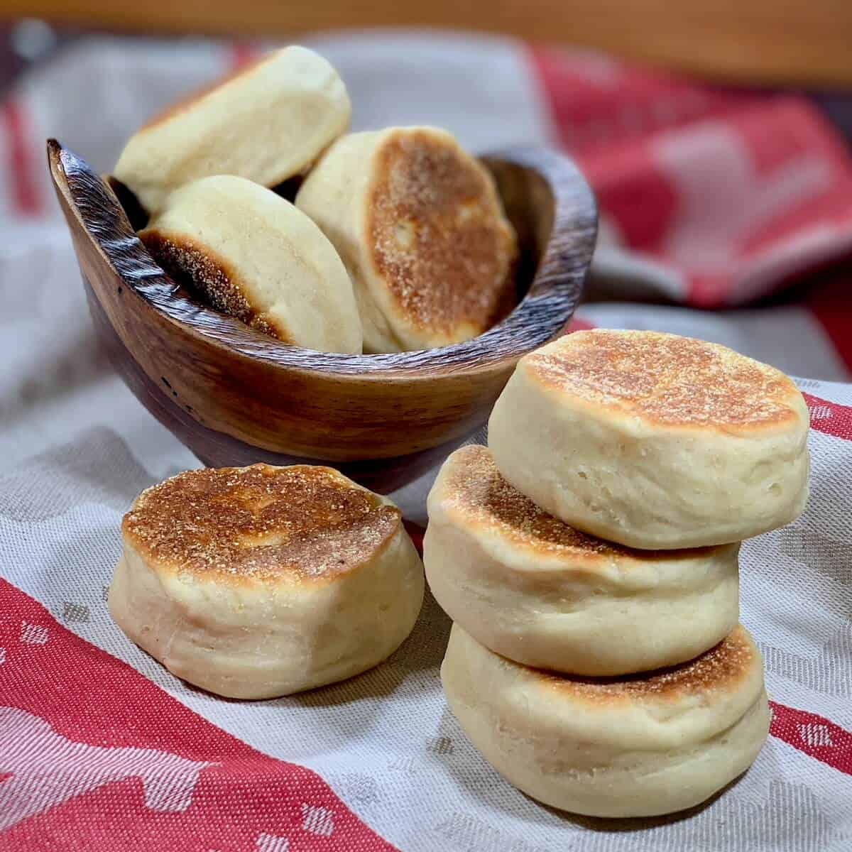 Three stacked Sourdough English Muffins with more in a wooden bowl on a red and white towel.