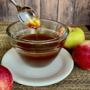 Boiled apple cider dripping from a spoon into a glass bowl on a white plate with apples.