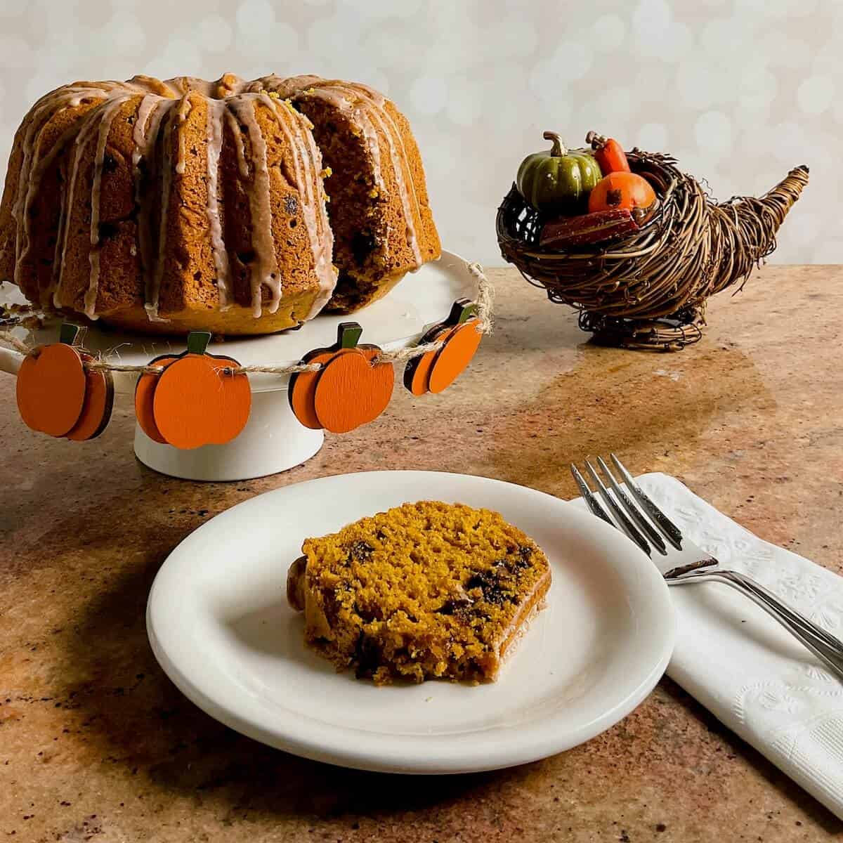 sourdough pumpkin cake on cake stand surrounded by plated slice & small pumpkins.