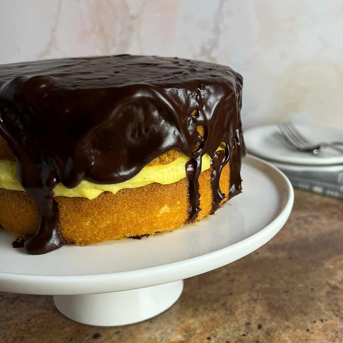 Boston Cream Pie on a white cake stand with plates & forks behind.