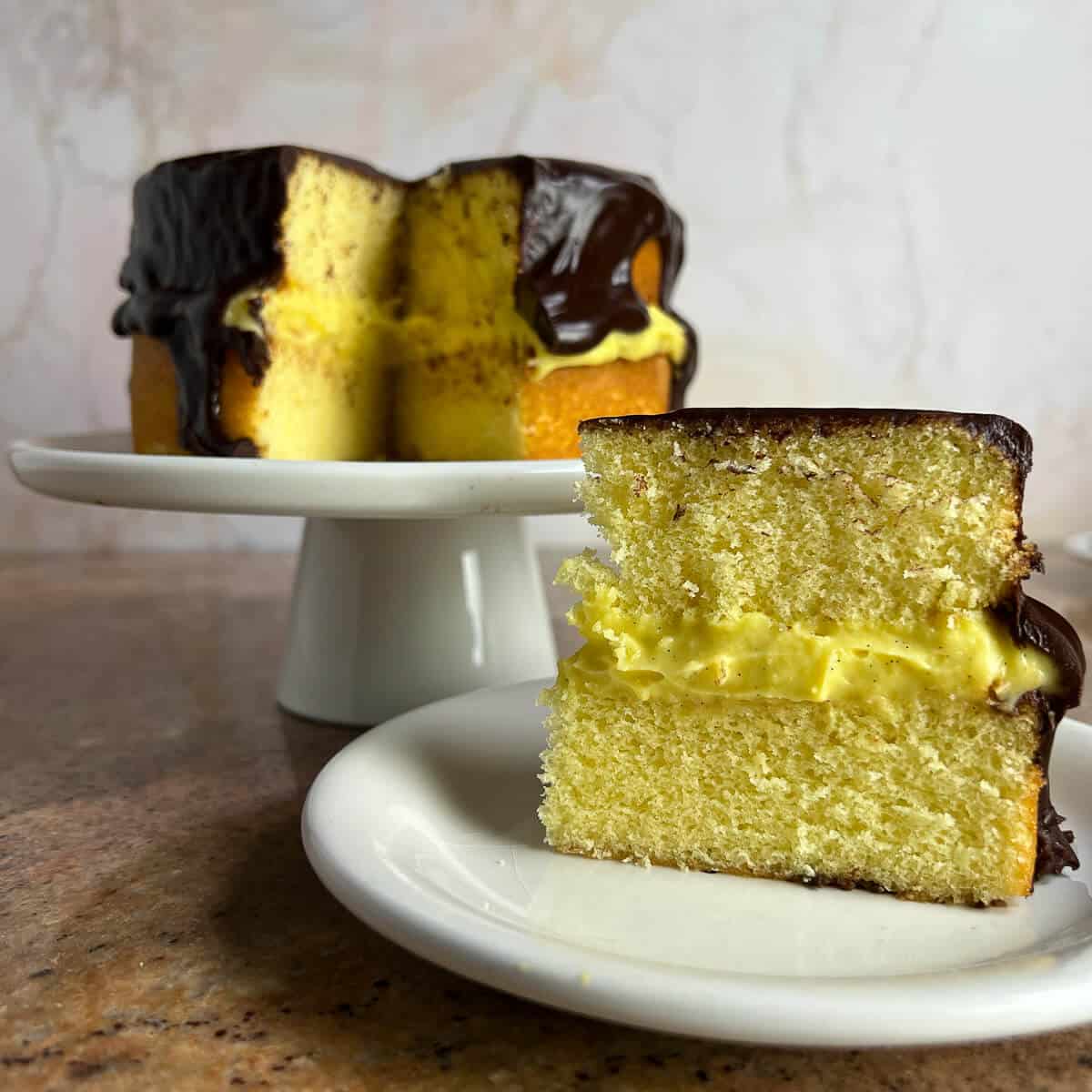 Slice of Boston Cream Pie on a white plate with the sliced whole cake on a white cake stand behind.