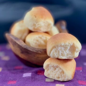 Two sourdough dinner rolls stacked on a purple checked towel with bowl of rolls in background.