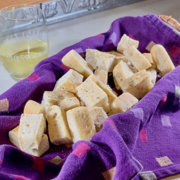 Sourdough Focaccia piled in purple towel lined basket next to a wine glass.