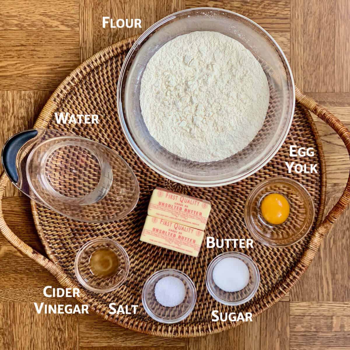 Short crust pastry ingredients portioned into glass bowls on a woven tray from overhead.