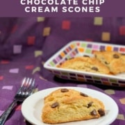 Chocolate chip cream scone on a white plate with a fork & more on a colorful plate behind all over a purple checked towel Pinterest banner.