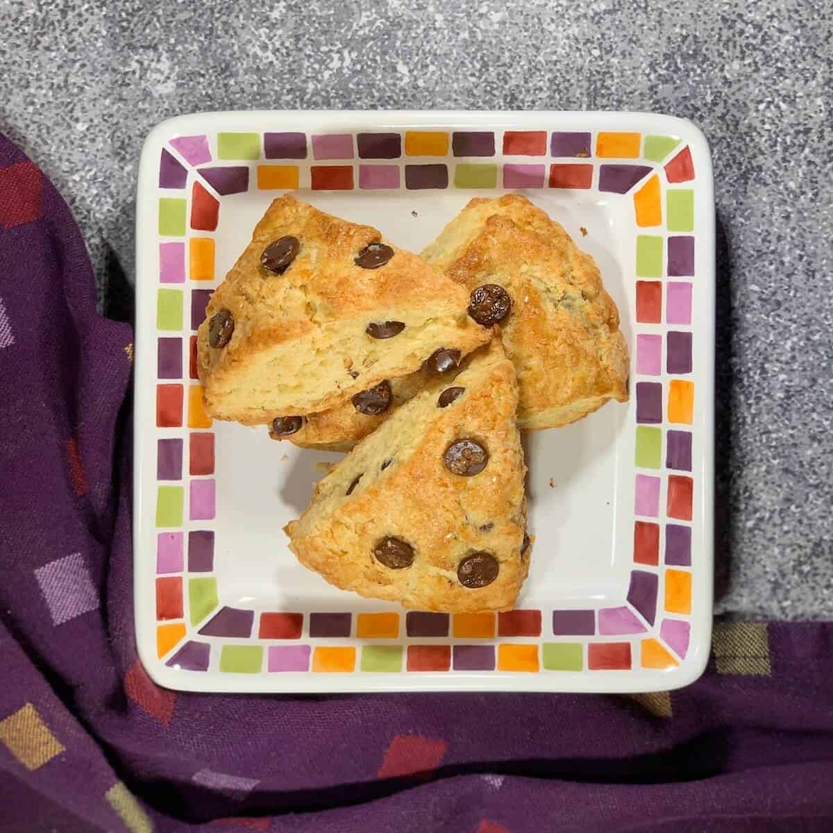 Chocolate chip cream scone on a white plate with a fork & more on a colorful plate behind all over a purple checked towel from overhead.