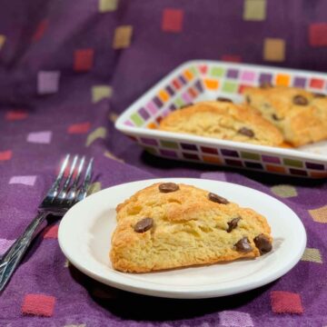 Chocolate chip cream scone on a white plate with a fork & more on a colorful plate behind all over a purple checked towel.