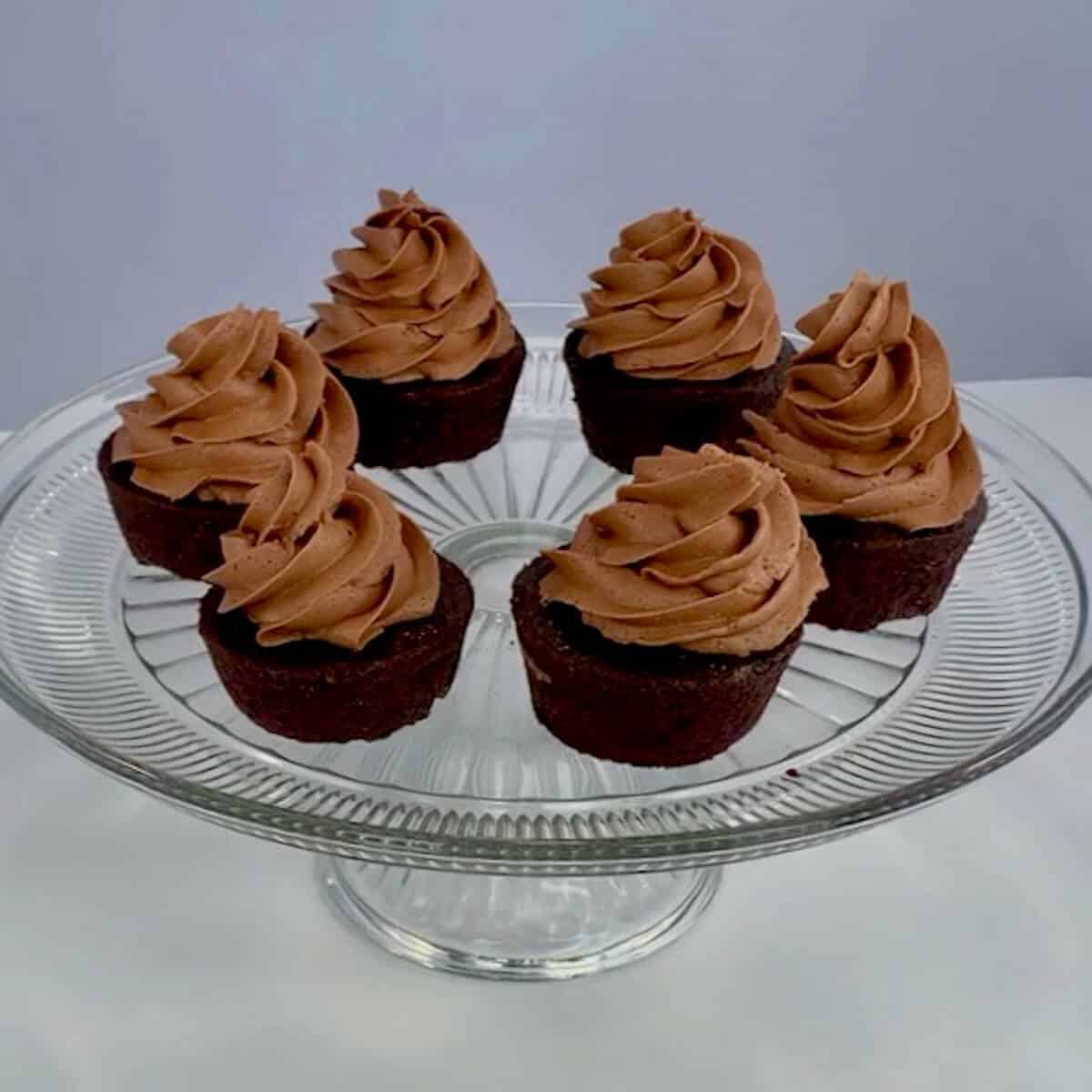 Six frosted chocolate cupcakes on a glass cake stand.