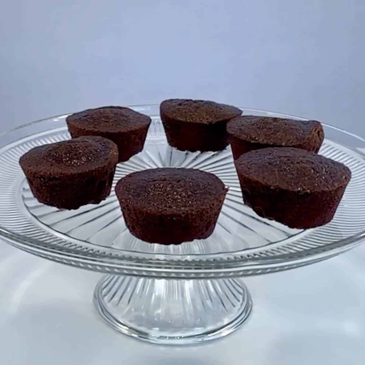 Six plain chocolate cupcakes on a glass cake stand.