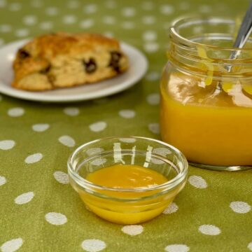 Lemon curd in a bowl and jar next to a scone on a white plate.