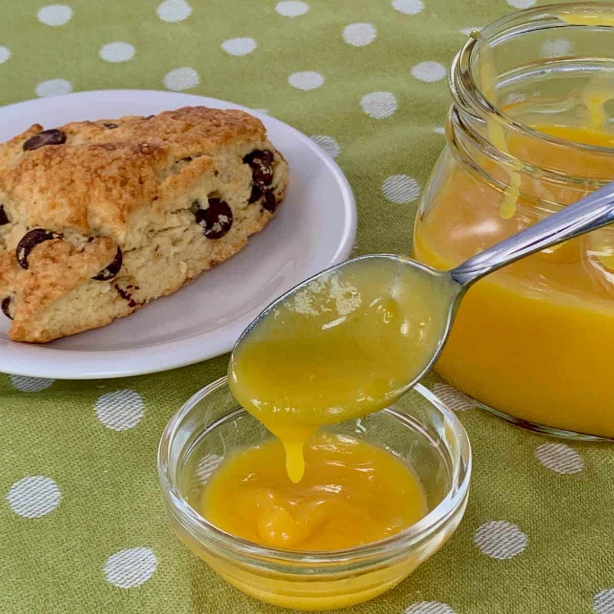 Lemon curd dripping from a spoon into a bowl and jar next to a scone on a white plate.