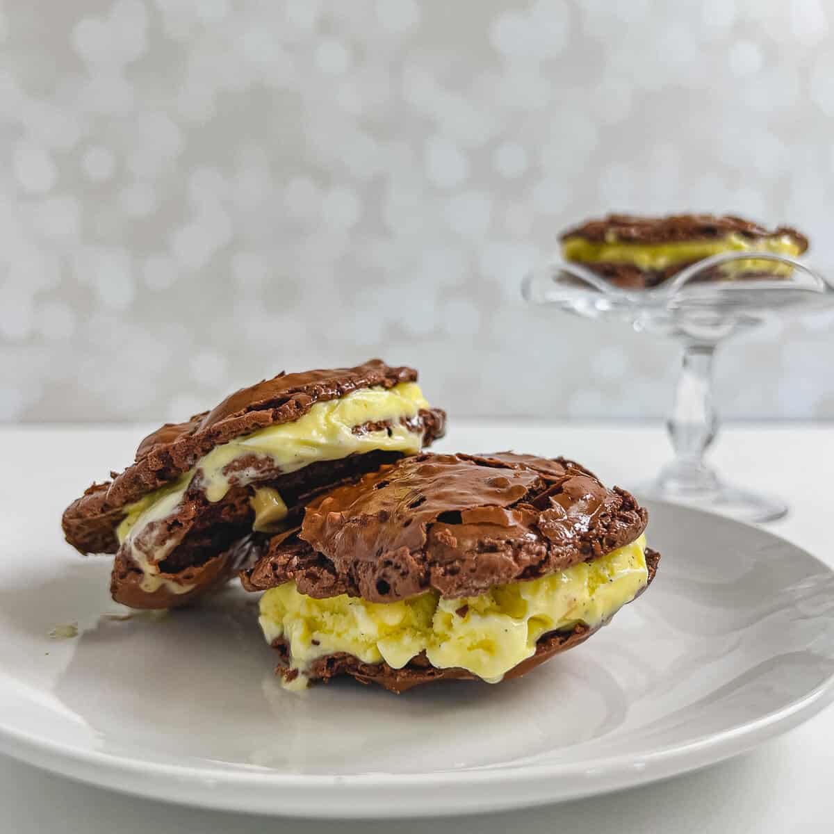 Two Gluten-Free Ice Cream Sandwiches stacked white plate with one on a glass cake stand behind.