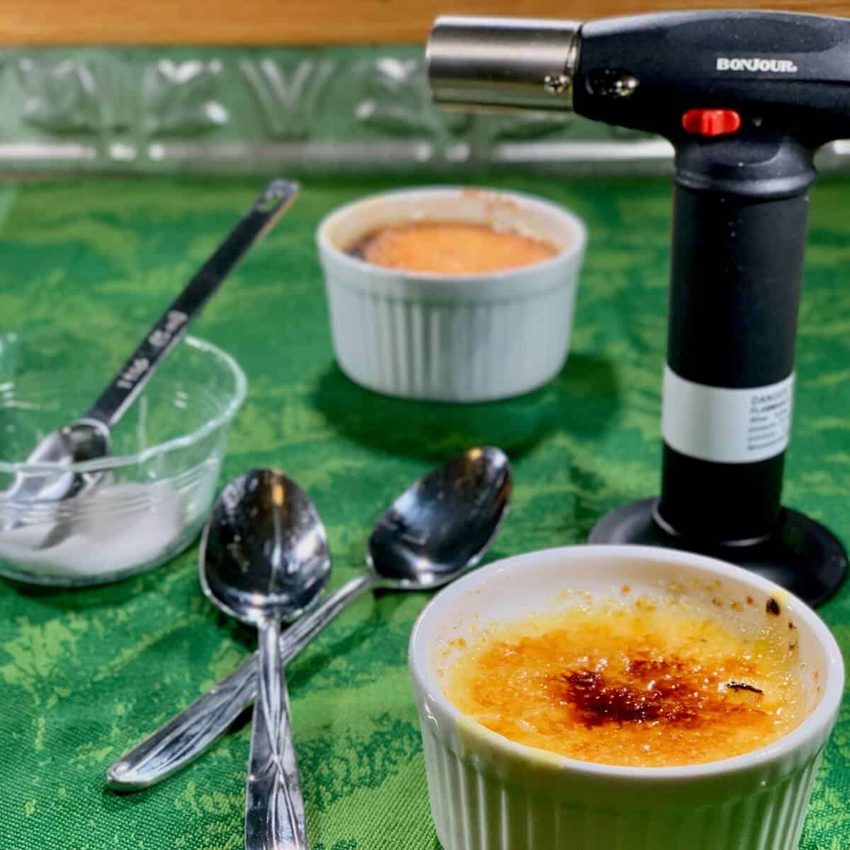 Creme Brulee on a green towel with torch, spoons, and sugar in background.