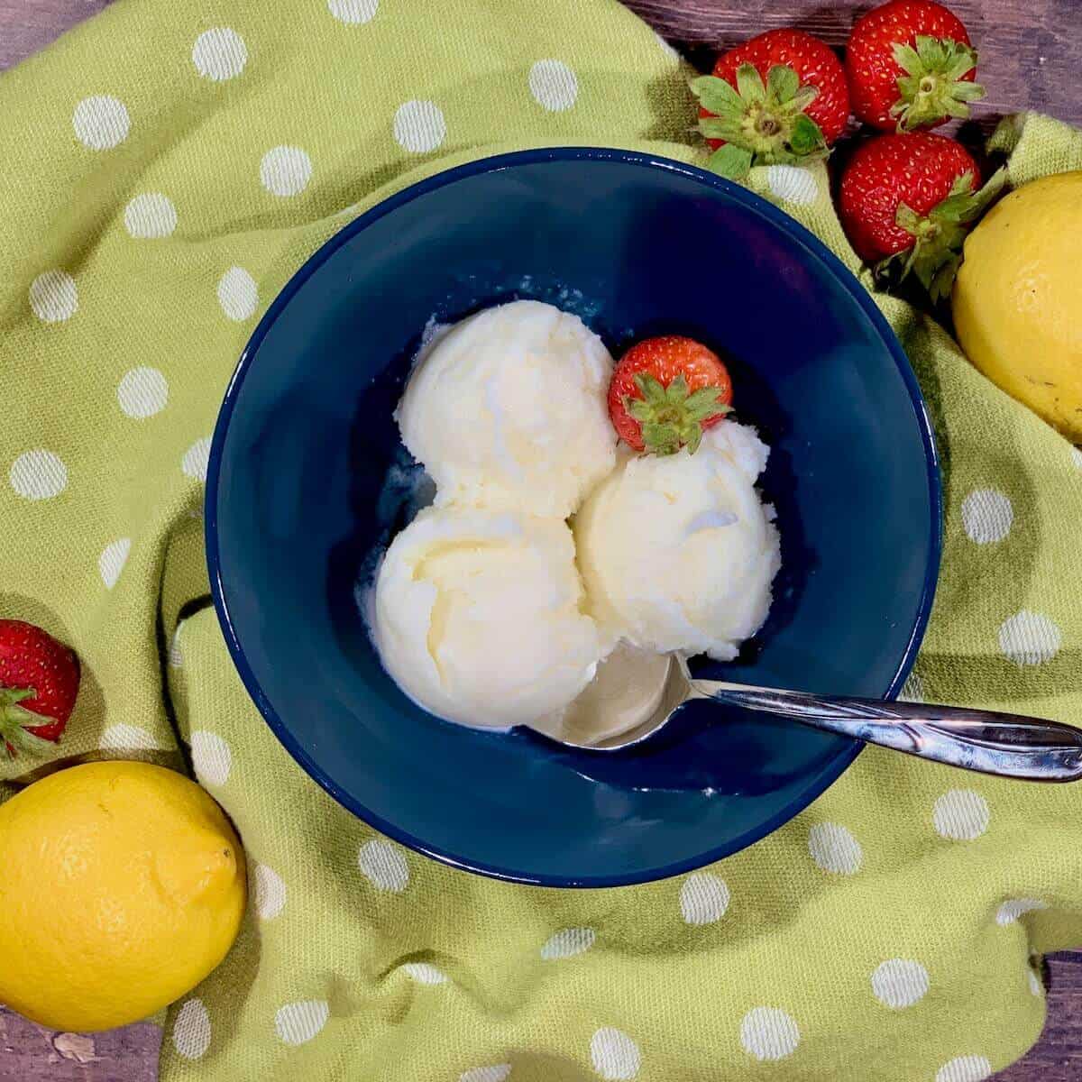 Lemon Sherbet scooped in blue bowl on green towel with strawberries & lemons from overhead.