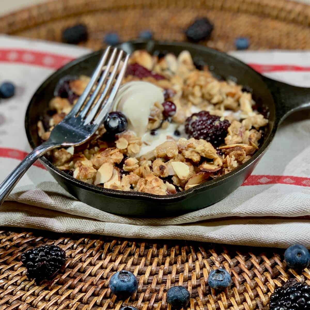 Mini Skillet Berry Crisp on a red & white striped towel with fork & scoop of vanilla ice cream surrounded by blueberries on a wooden tray.
