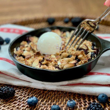 Mini Skillet Berry Crisp on a red & white striped towel with fork lifted & scoop of vanilla ice cream surrounded by blueberries on a wooden tray.