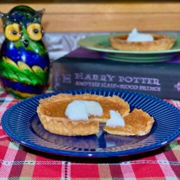 Treacle Tart sliced & plated on blue plate with tart on green plate on book & glass owl in background.