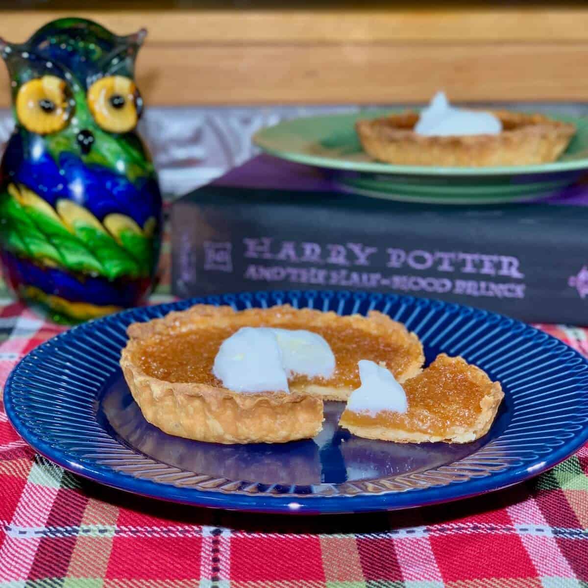 Treacle Tart sliced & plated on blue plate with tart on green plate on book & glass owl in background.