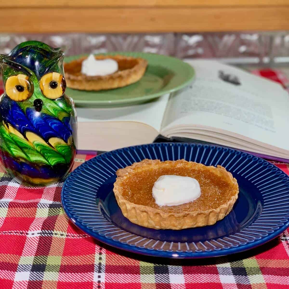 Treacle Tart on blue plate with tart on green plate on open book & glass owl in background.