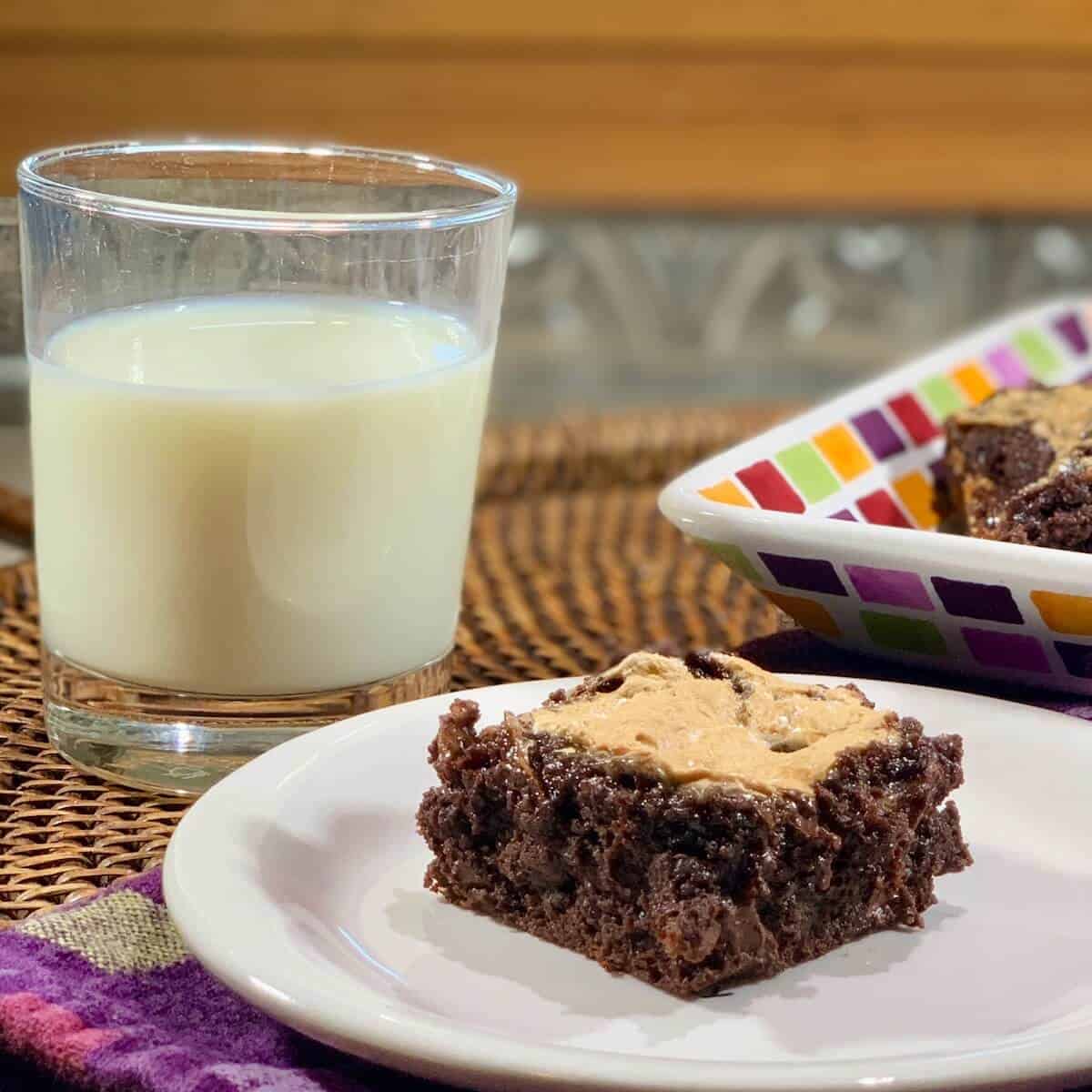 Marshmallow topped brownie on a white plate next to a glass of milk all on a woven tray.