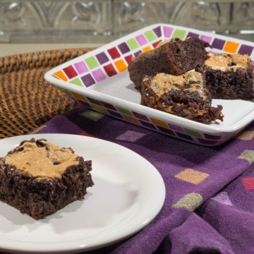 Marshmallow topped brownie on a white plate with more on a multicolored checked plate behind on a purple checked towel.