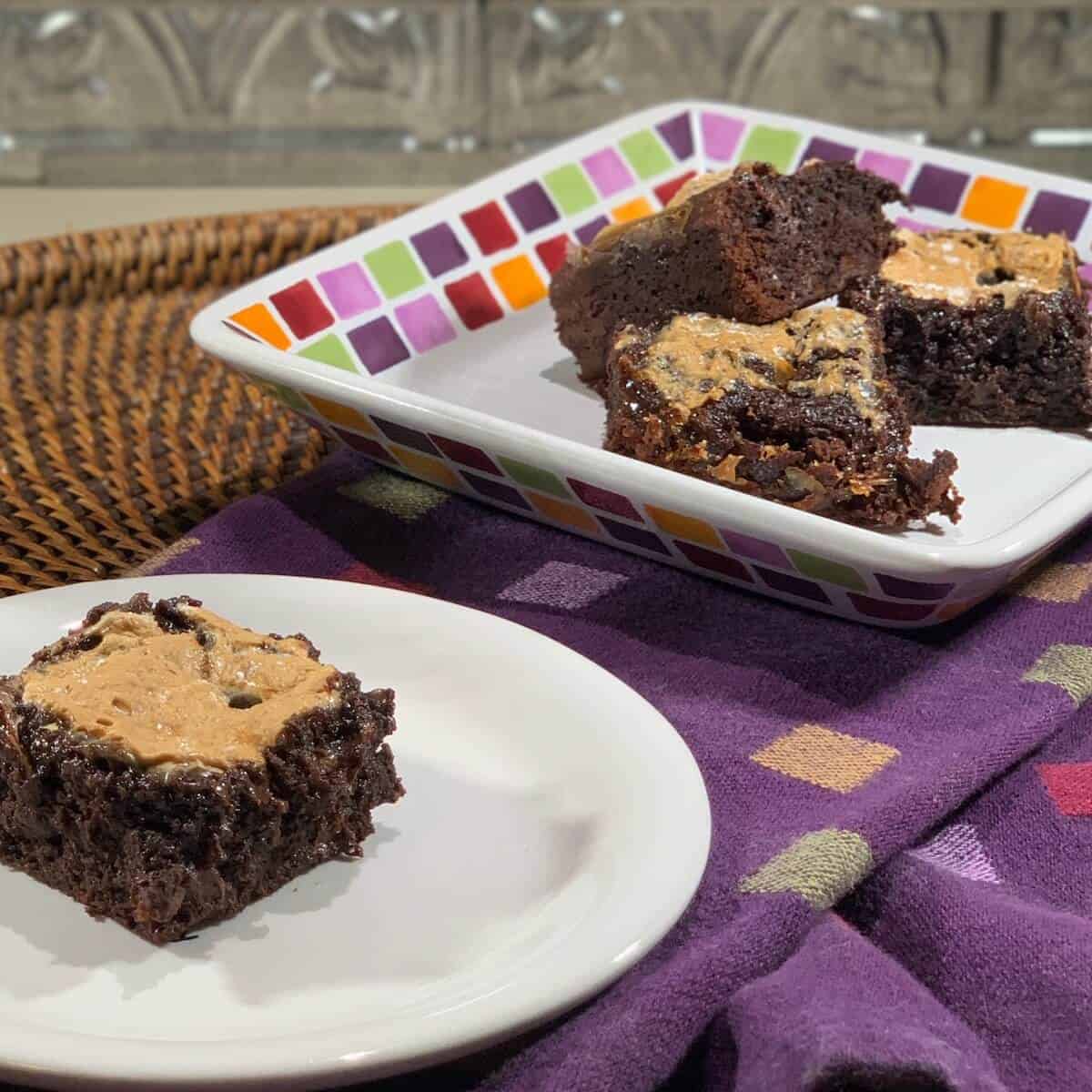 Marshmallow topped brownie on a white plate with more brownies on a multicolored checked plate behind on a purple checked towel.