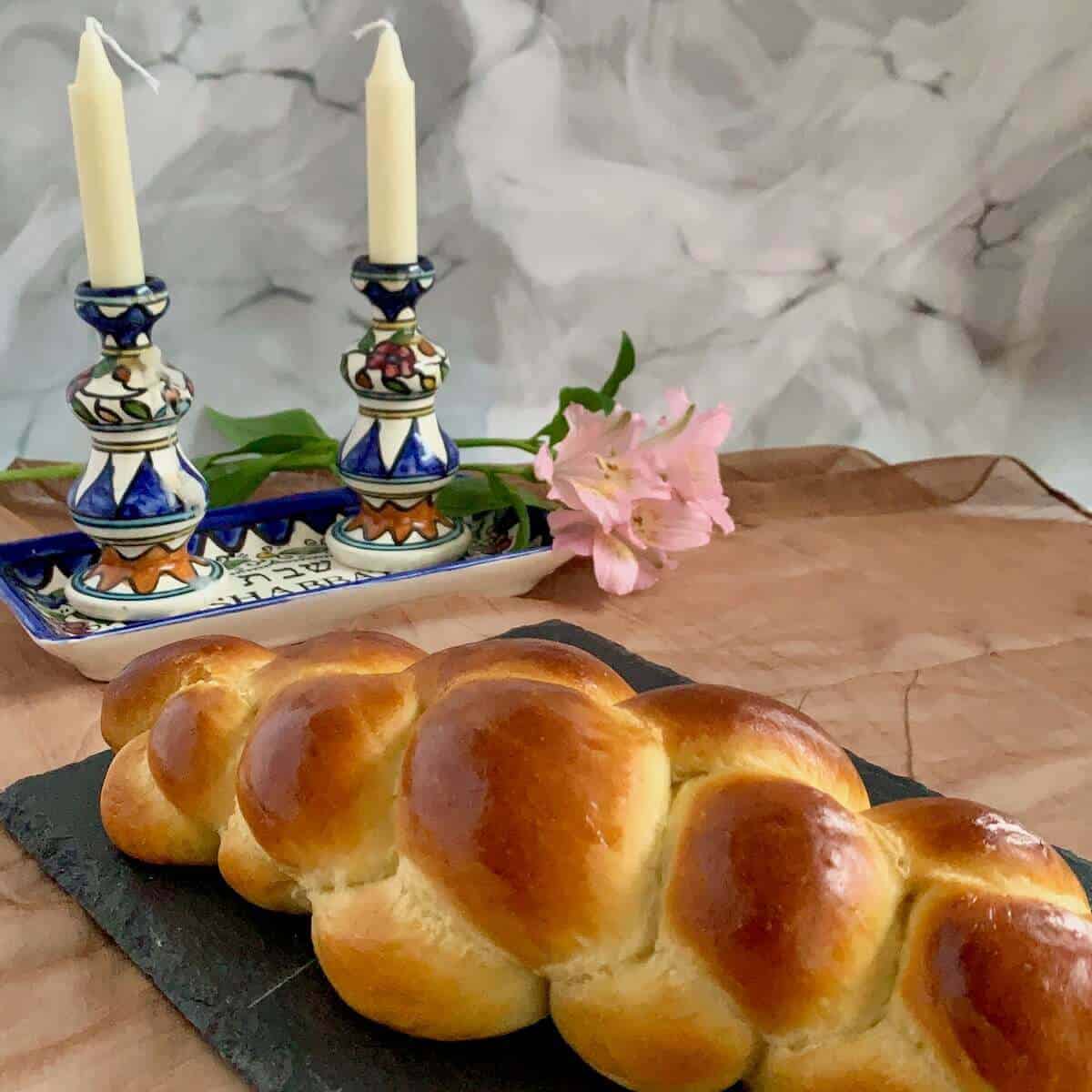 Oblong challah on a black slate with Shabbat candles in the background.