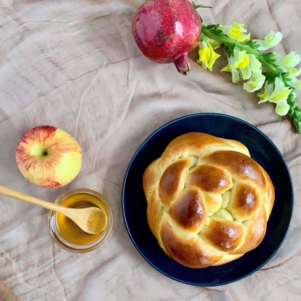 Round challah on a plate with apple, honey, & pomegranate viewed from overhead.