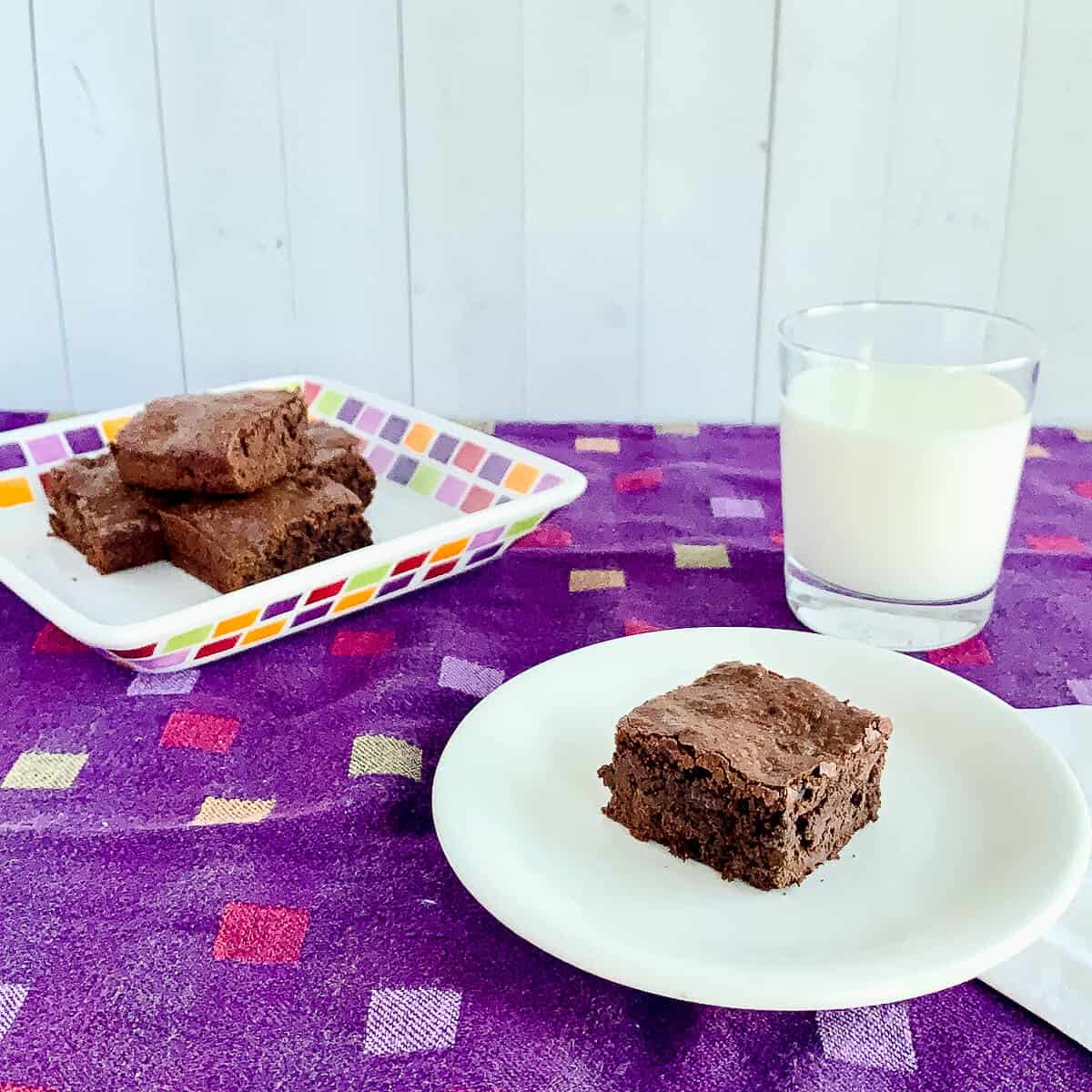 Cake brownie on white plate with a stack of brownies & milk in background.