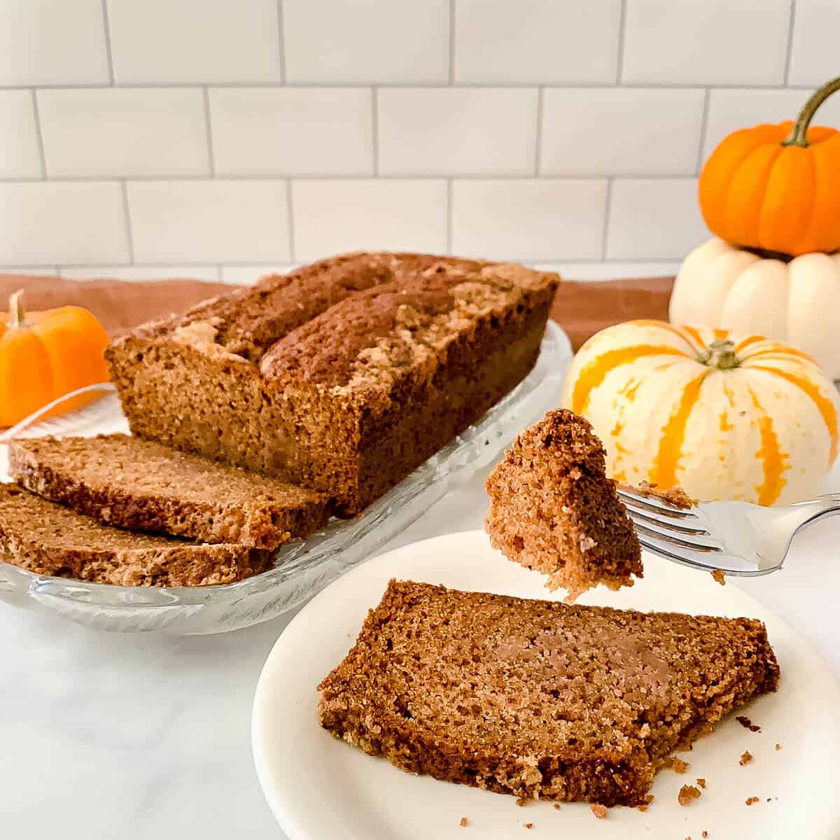 Sourdough Spice Cake slice on white plate with bite on a fork with loaf & pumpkins behind.