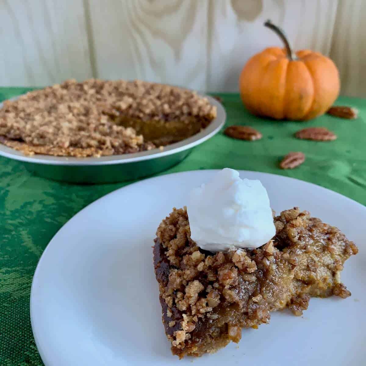 Praline Pumpkin Pie in pie pan with a slice topped with whipped cream on a white plate next to pumpkin & scattered pecans.