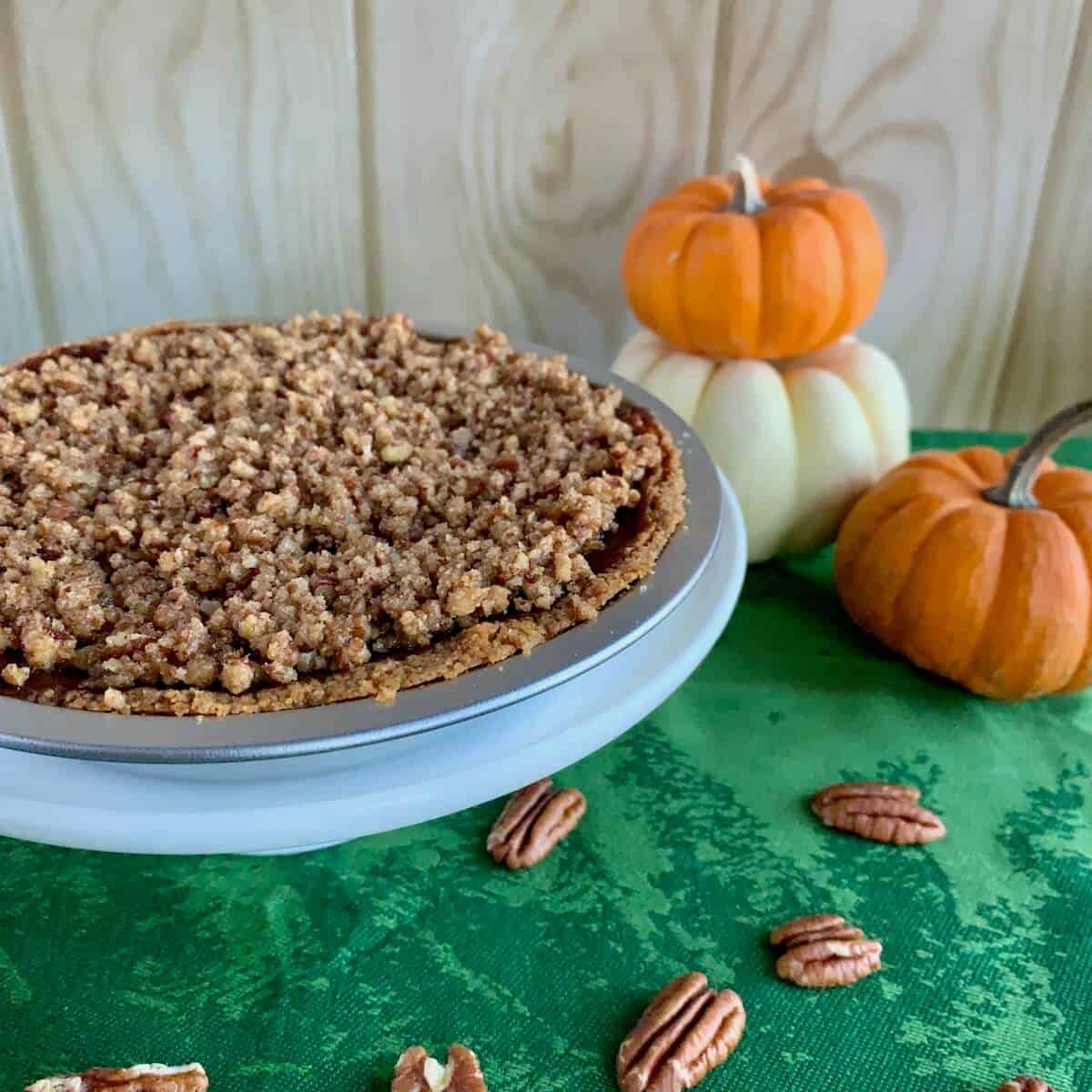 Whole Praline Pumpkin Pie on a white cake stand with a stack of pumpkins behind & scattered pecans.