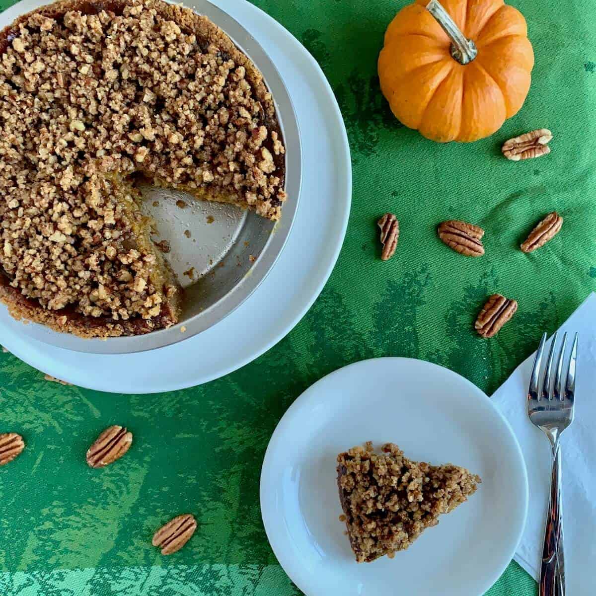 Praline Pumpkin Pie in pie pan with a slice on a white plate next to pumpkin & scattered pecans from overhead.